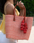 Model wearing Cancun Small Sisal Basket Bag with Tassel Charm handmade natural sisal fibers woven into even width horizontal bands of coral orange and natural straw color that create a pinstripe pattern with matching knotted raffia tufts tassel charm embellishment and leather handles beach bag - Shebob