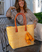 Model wearing Cancun Small Sisal Basket Bag with Tassel Charm handmade natural sisal fibers woven into even width horizontal bands of saffron yellow and natural straw color that create a pinstripe pattern with matching knotted raffia tufts tassel charm embellishment and leather handles beach bag - Shebob