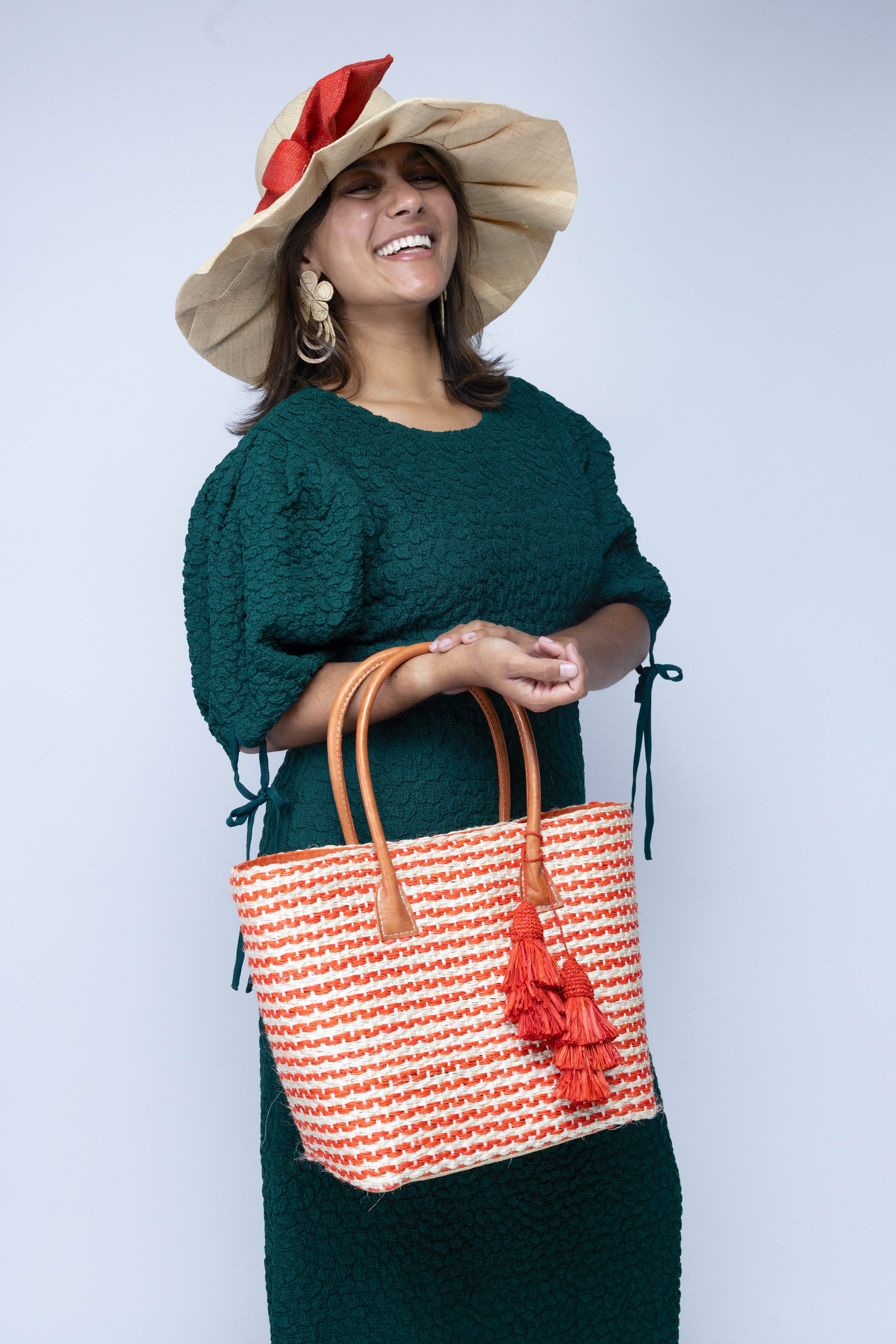 Model wearing Cancun Small Sisal Basket Bag with Tassel Charm handmade natural sisal fibers woven into even width horizontal bands of coral orange and natural straw color that create a pinstripe pattern with matching knotted raffia tufts tassel charm embellishment and leather handles beach bag - Shebobo (with Lilly 5" Wide Brim Pleated Fan Straw Sun Hat with Big Bow)