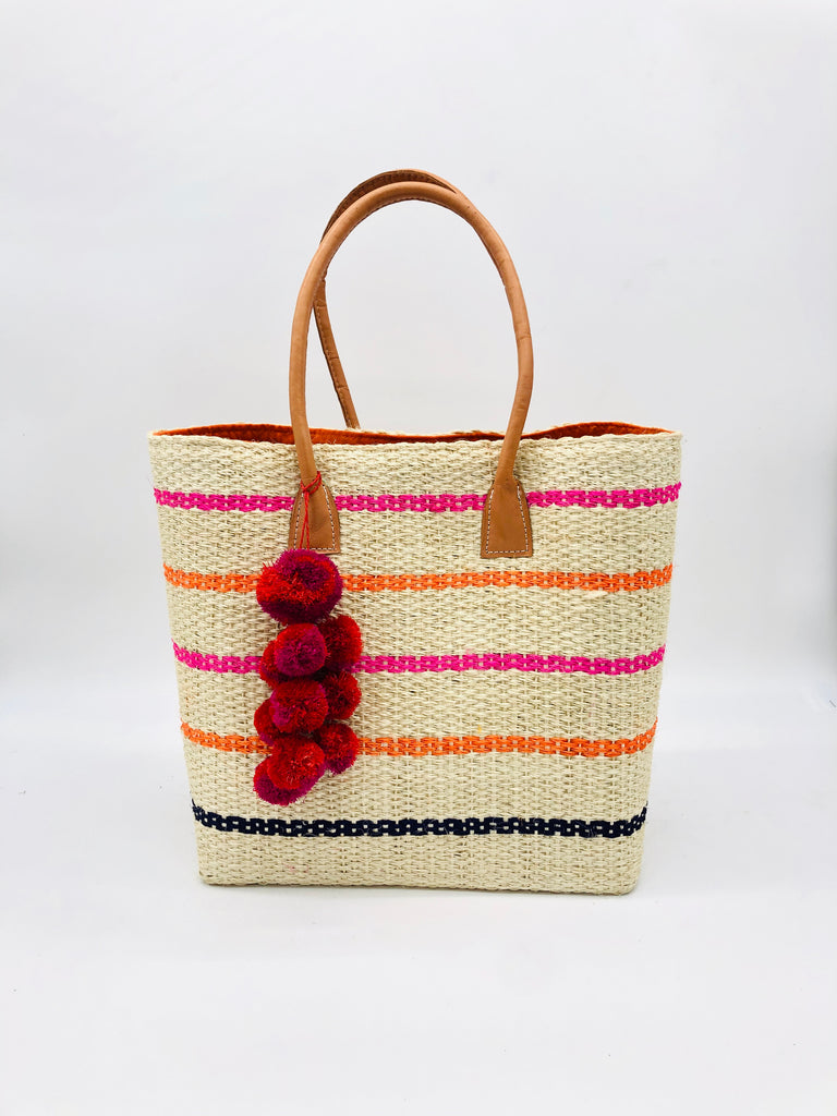Capitola Pink Multi Pinstripes Sisal Basket Bag with Waterfall Pompoms handmade woven natural sisal fiber tote bag in wide bancs of natural straw color with narrow bands of fuchsia pink, coral orange/red, and navy blues in a horizontal stripe pattern with leather handles and matching waterfall pompom charm embellishment - Shebobo