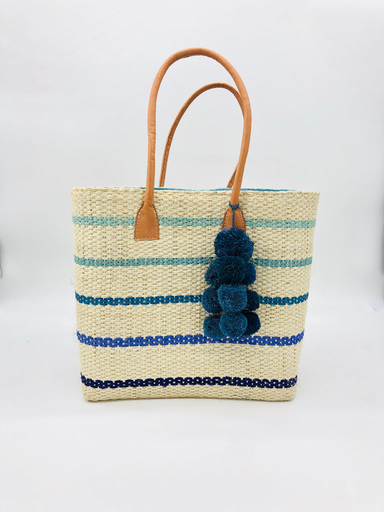 Capitola Turquoise Multi Pinstripes Sisal Basket Bag with Waterfall Pompoms handmade woven natural sisal fiber tote bag in wide bancs of natural straw color with narrow bands of light blue, seafoam blue/green, turquoise, royal, and navy blues in a horizontal stripe pattern with leather handles and matching waterfall pompom charm embellishment - Shebobo
