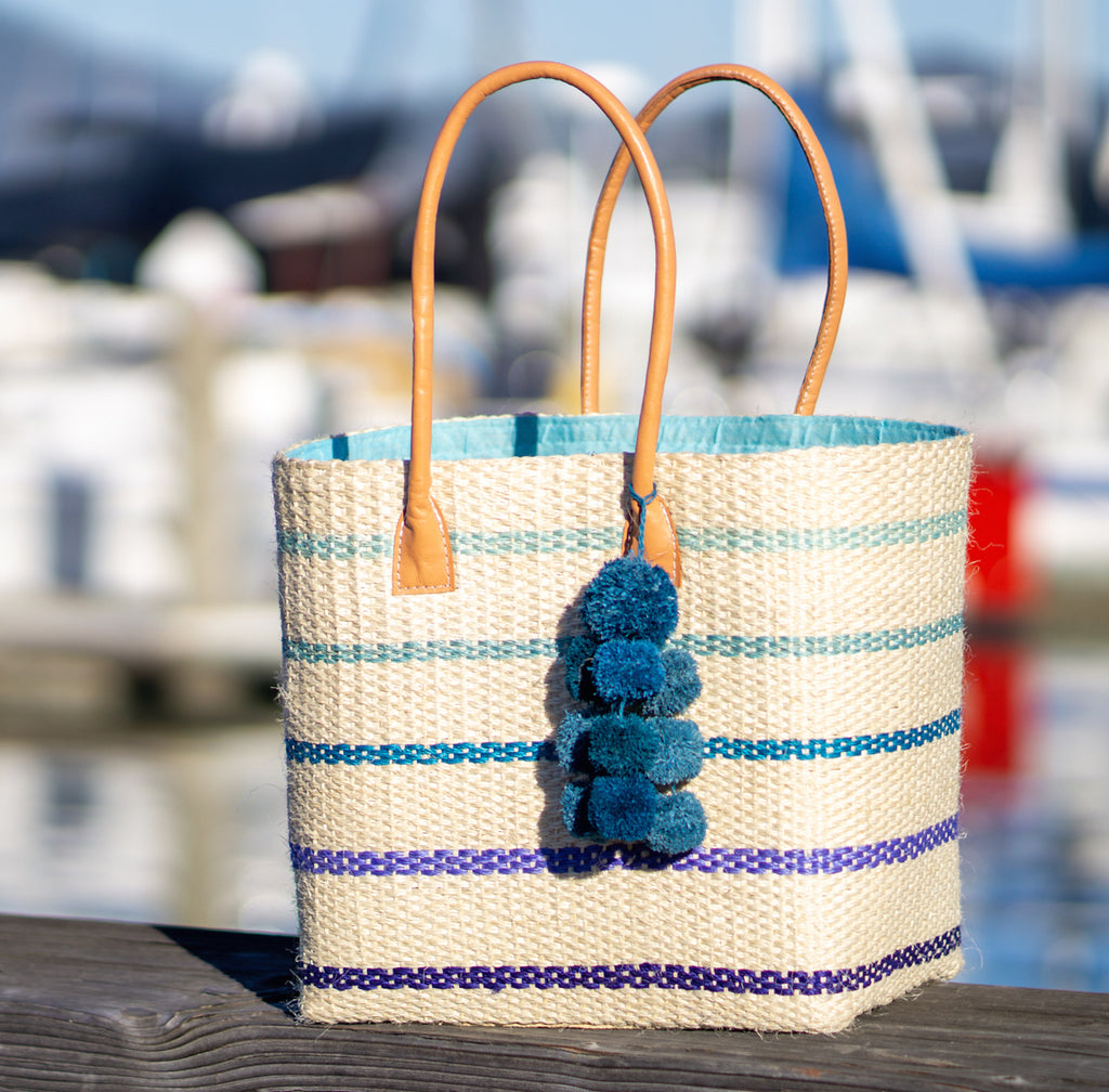 Lifestyle shot of Capitola Turquoise Multi Pinstripes Sisal Basket Bag with Waterfall Pompoms handmade woven natural sisal fiber tote bag in wide bancs of natural straw color with narrow bands of light blue, seafoam blue/green, turquoise, royal, and navy blues in a horizontal stripe pattern with leather handles and matching waterfall pompom charm embellishment - Shebobo