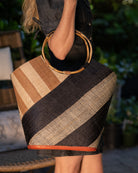 Model wearing Carmen Double Wide Stripe Black & Cappuccino Straw Bucket Bag w/ Bamboo Handle handmade loomed raffia palm fiber in even width wide stripe pattern of black and natural straw color oriented on the diagonal of three quarters of the bag with matching stripe pattern of cappuccino tea/light/brown and natural diagonally on the top left corner quarter shoulder bag handbag - Shebobo