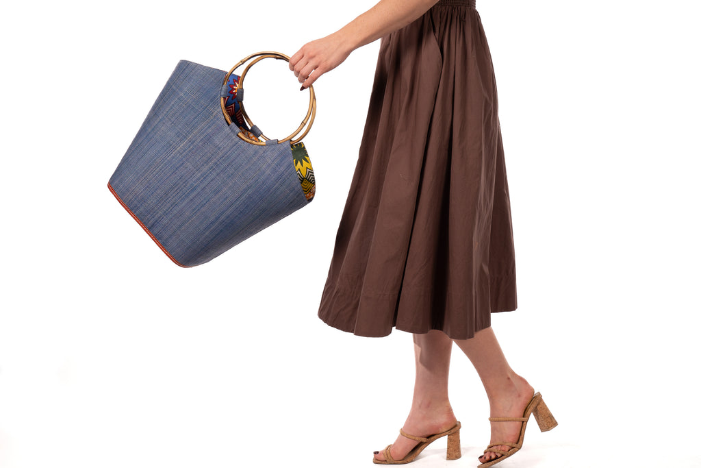 Model wearing Carmen Denim Straw Bucket Bag with Bamboo Handles handmade loomed raffia in a solid hue of denim/retro/light/blue with assorted African print fabric liner handbag - Shebobo