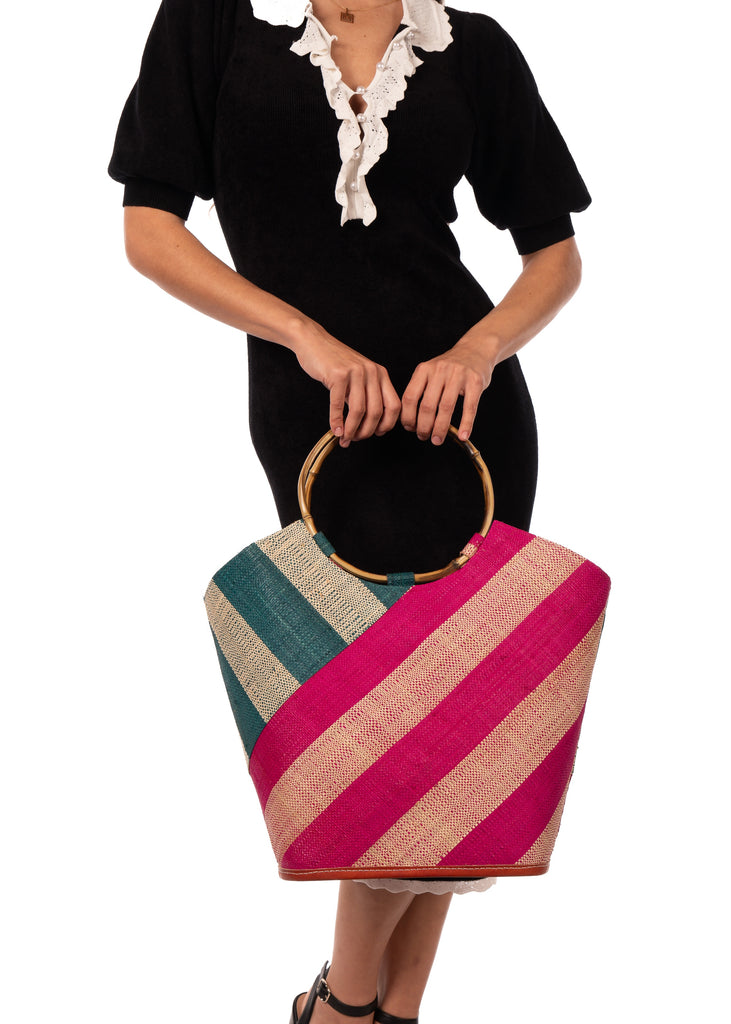 Model wearing Carmen Double Wide Stripe Fuchsia & Teal Straw Bucket Bag w/ Bamboo Handle handmade loomed raffia palm fiber in even width wide stripe pattern of Fuchsia hot/bright/barbie/pink and natural straw color oriented on the diagonal of three quarters of the bag with matching stripe pattern of teal dark/blue/green and natural diagonally on the top left corner quarter shoulder bag handbag - Shebobo