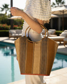 Model wearing Carmen Straw Bucket Bag with Bamboo Handles Cinnamon Swirl multicolor vertical stripe pattern of natural straw color, cinnamon/tobacco/dark/brown, and grey with assorted liner purse - Shebobo