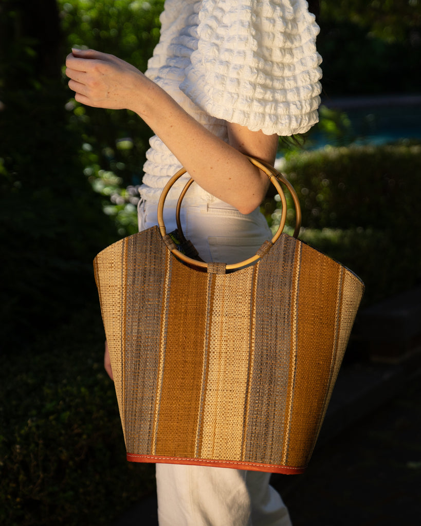 Model wearing Carmen Straw Bucket Bag with Bamboo Handles Cinnamon Swirl multicolor vertical stripe pattern of natural straw color, cinnamon/tobacco/dark/brown, and grey with assorted liner purse - Shebobo