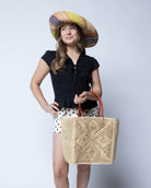 Model wearing Cheval Lace Square Pattern Crochet Straw Basket Bag handmade from natural raffia palm fiber in a lace square geometric diamond pattern with triangle detailing textile in a natural straw color with leather handles handbag purse beach bag tote- Shebobo (with Monica 5" & 7" Wide Brim Multicolor Stripe Pattern Packable Straw Sun Hats)