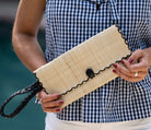 Model wearing ChiChi Natural Straw Clutch Bag handmade loomed raffia palm fiber wristlet in natural straw color with black cross stitch edging, button & loop closure, and braided wrist strap - Shebobo