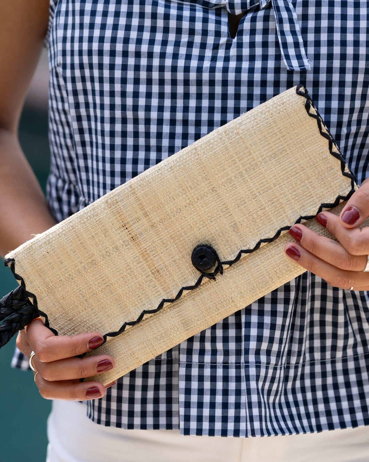 Model wearing ChiChi Natural Straw Clutch Bag handmade loomed raffia palm fiber wristlet in natural straw color with black cross stitch edging, button & loop closure, and braided wrist strap - Shebobo