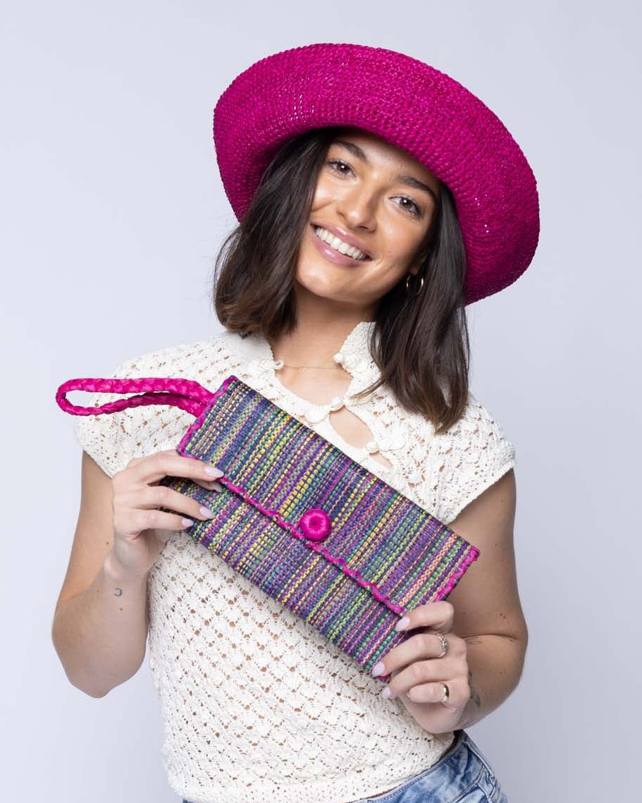 Model wearing ChiChi Multicolor Melange Multicolor Straw Clutch Bag handmade loomed raffia palm fiber wristlet in vertical heathered/blended stripe pattern of orchid pink/purple, navy dark/blue, fuchsia bright/pink, yellow, midori lime/green, light pink, etc. with matching color cross stitch edging, button & loop closure, and braided wrist strap - Shebobo