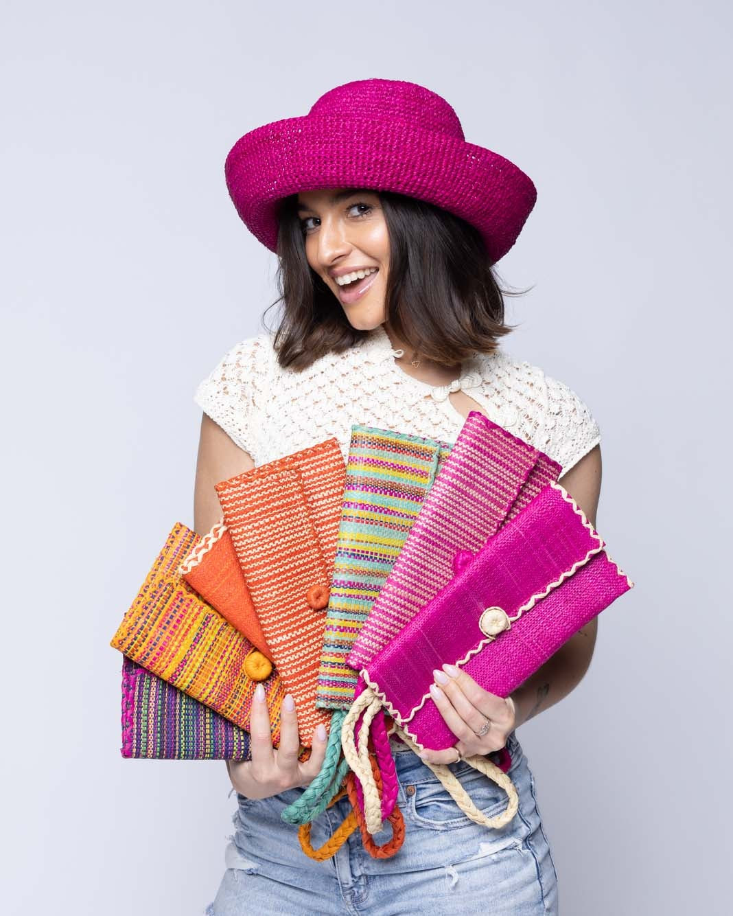 Model wearing multiple solid hues, two tone stripes, multicolor stripes, and melange patterns of ChiChi Straw Clutch Wristlet Bag handmade loomed raffia palm fiber in vibrant colors with natural cross stitch edging and raffia loop button closure with matching braided wrist loop  - Shebobo