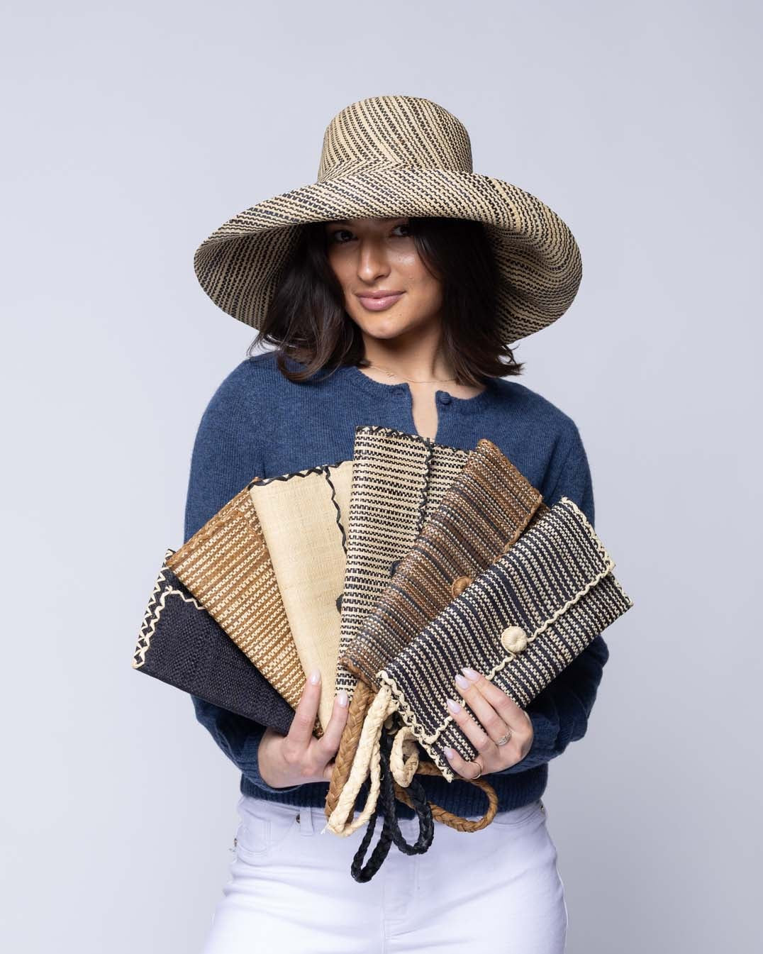 Model wearing multiple solid hues, two tone stripes, multicolor stripes, and melange patterns of ChiChi Straw Clutch Wristlet Bag handmade loomed raffia palm fiber in vibrant colors with natural cross stitch edging and raffia loop button closure with matching braided wrist loop  - Shebobo