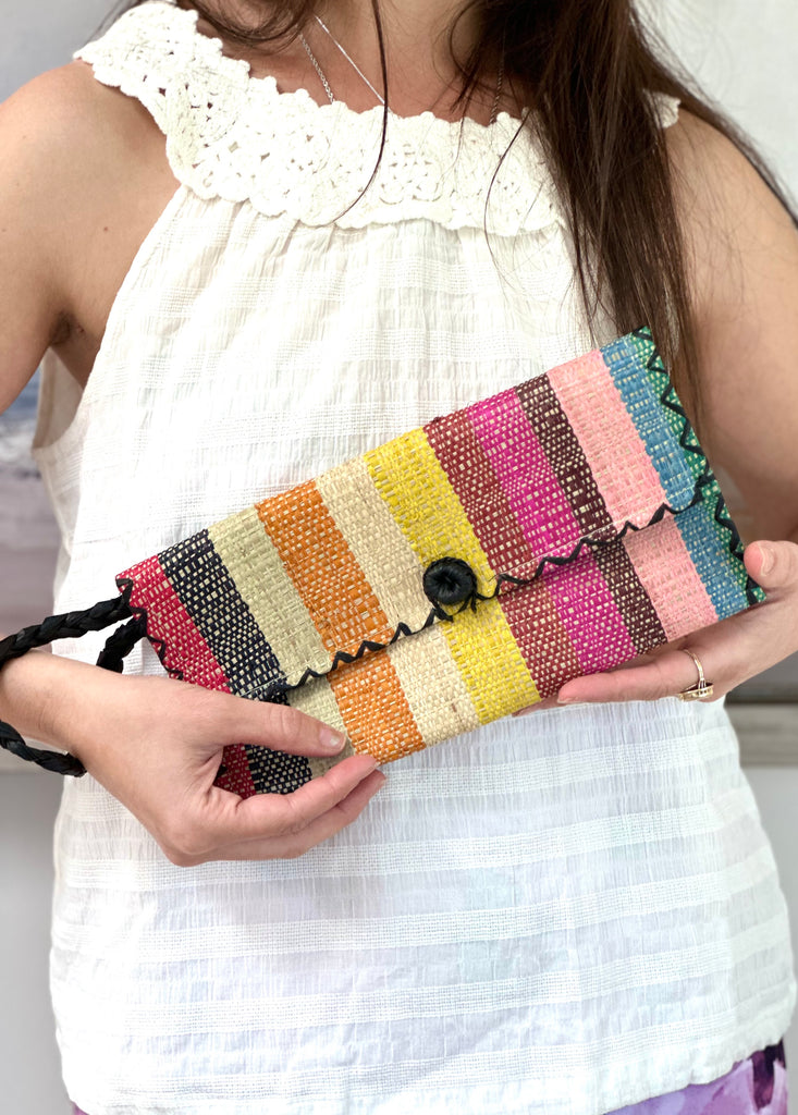 Model wearing ChiChi Lollipop Stripe Multicolor Straw Clutch Bag handmade loomed raffia palm fiber wristlet in multi-width bands of turquoise blue, saffron yellow, blue, natural, fuchsia pink, green, black, coral orange/red, etc. that create a vertical stripe pattern with black color cross stitch edging, button & loop closure, and braided wrist strap - Shebobo