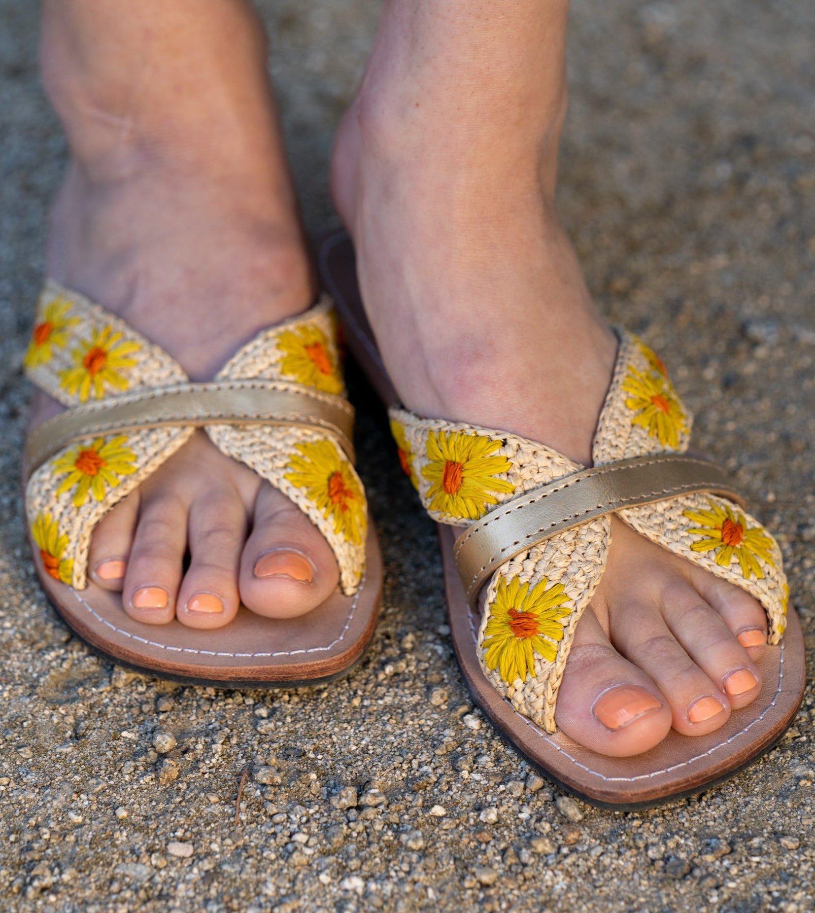 Model wearing Daisy Crochet Straw & Leather Sandal with Flower Embroidery & Metallic Detailing handmade crochet natural raffia palm fiber in a solid hue of natural straw color in two bands with saffron yellow petals and coral orange/red centered embroidered flower pattern wrap around the top  of the shoe with a band of metallic gold across the top summer sandals shoes - Shebobo
