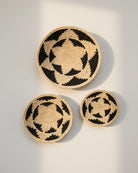 Life style view Dakarai Raffia Straw Wall Decor Display Basket Tray: Set of 3 handmade woven natural raffia palm fiber in a crochet loop technique concentric coiled circular pattern with double natural straw color and black centered star flower pattern set of three sized flat bowl trays - Shebobo