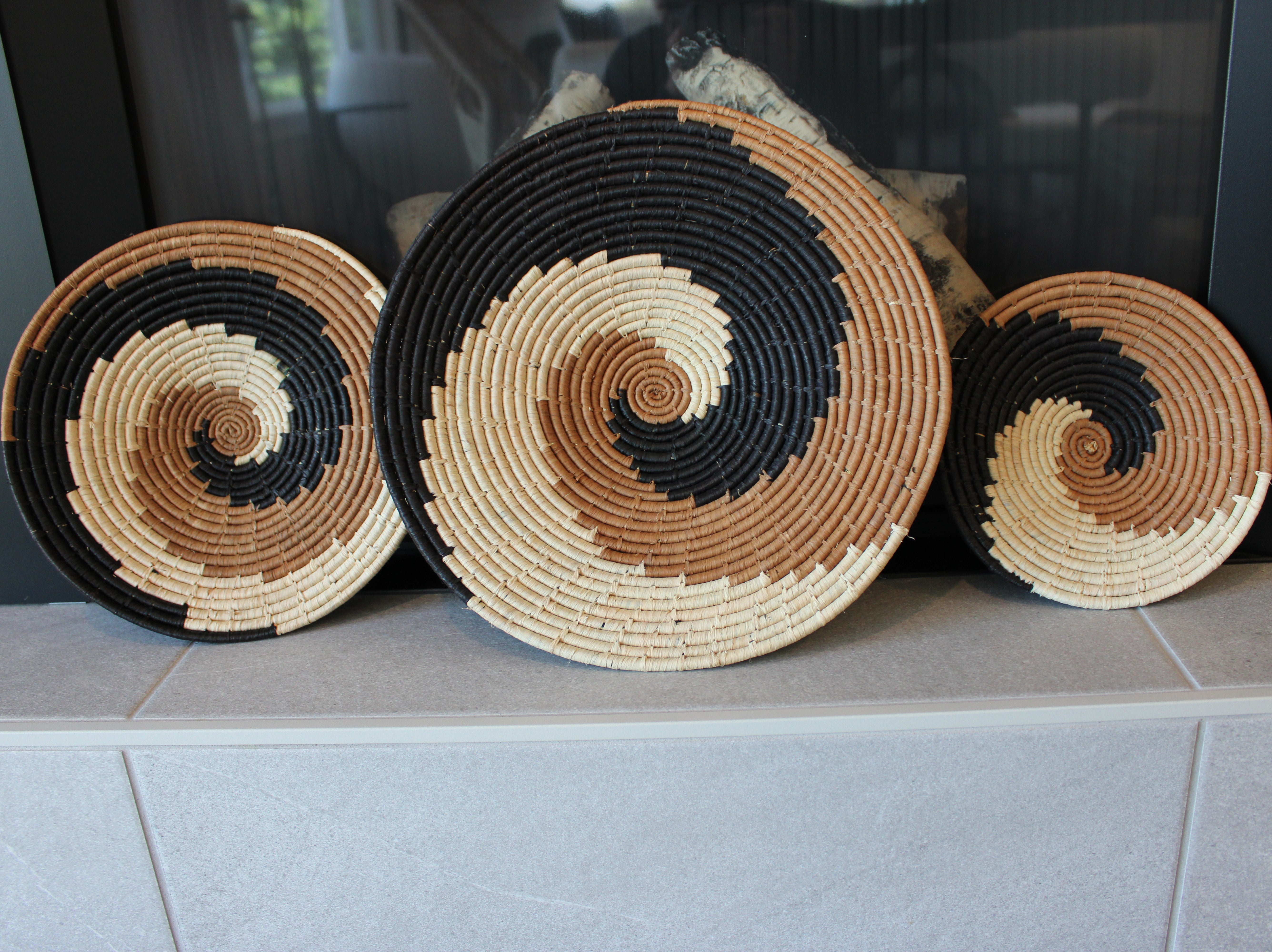 Home decor lifestyle view Dakarai Raffia Straw Wall Decor Display Basket Trays: Set of 3 handmade woven raffia palm fiber in a crochet loop technique creating a concentric swirl pattern of bands of natural straw color, cinnamon/tobacco/brown, and black in a three tone spiral from the center to the outer edge set of three sized flat bowl trays - Shebobo