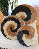 Home decor lifestyle view Dakarai Raffia Straw Wall Decor Display Basket Trays: Set of 3 handmade woven raffia palm fiber in a crochet loop technique creating a concentric swirl pattern of bands of natural straw color, cinnamon/tobacco/brown, and black in a three tone spiral from the center to the outer edge set of three sized flat bowl trays - Shebobo