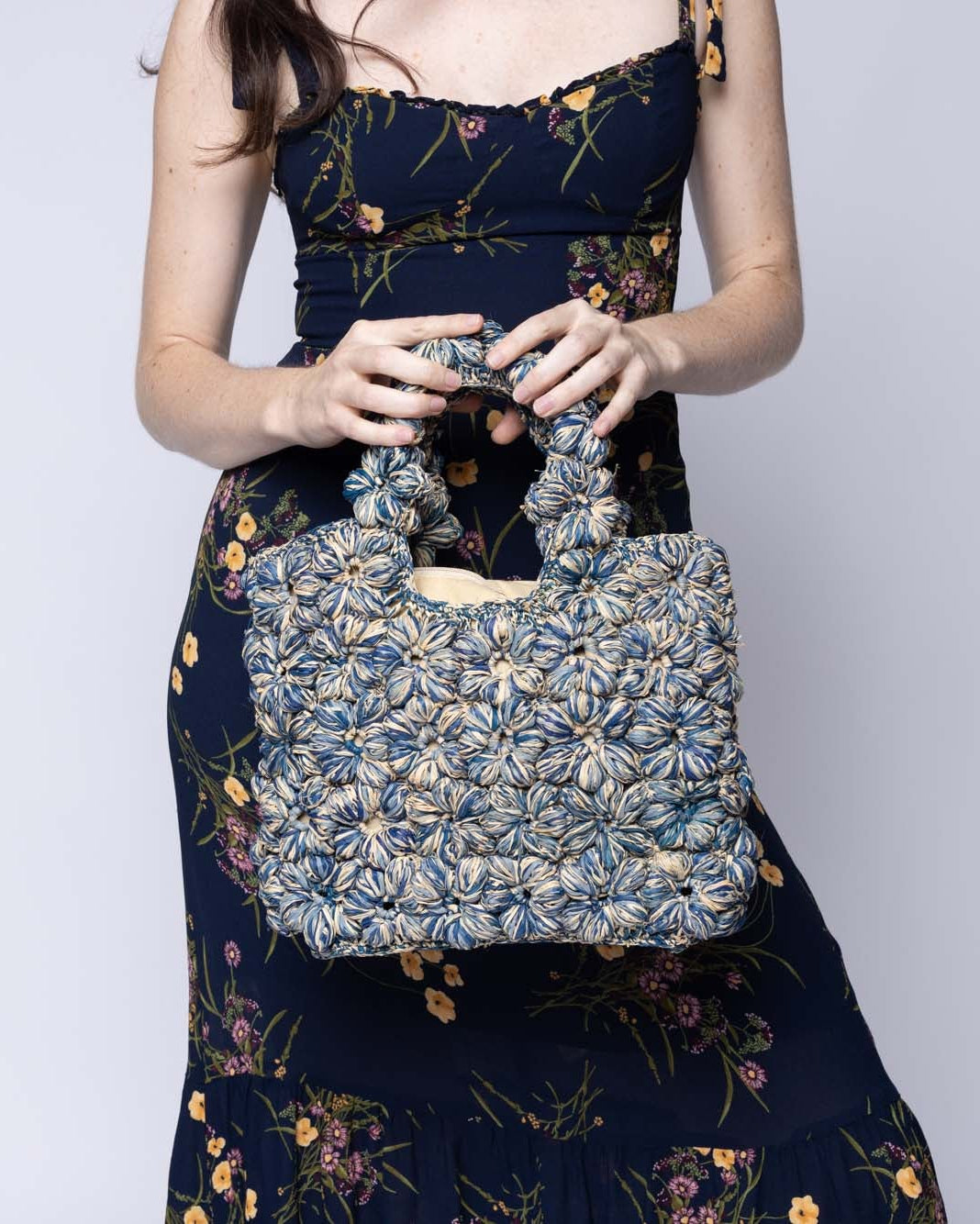 Model wearing Florence Denim Blues Multicolor Flower Crochet Straw Basket Handbag handmade natural raffia palm fiber in a multicolor hue combination of natural straw color, denim light/retro/blue, and luminous navy/dark/blue with subtle tied flower design consisting of 88 flowers crochet together along the outside of the bag as well as the handles purse tote - Shebobo