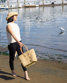 Model wearing Flower Power Natural Crochet Straw Basket Bag handmade from natural raffia palm fiber in a granny square-esque geometric floral pattern with flower detailing projecting from main crochet textile in a natural straw color with leather handles handbag purse - Shebobo (with Leor 3" Natural Crochet Straw Sun Hat)