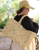 Model wearing Gemma Crochet Disc Rectangle Large Straw Basket Handbag handmade braided natural raffia palm fiber in a solid hue of Natural straw color with subtle square disc pattern around the outside of the bag in two horizontal bands of squares creating a geometric pattern and matching woven handles beach bag purse - Shebobo (with Rachel 4" Brim Crochet Straw Sun Hats with Colored Edge)