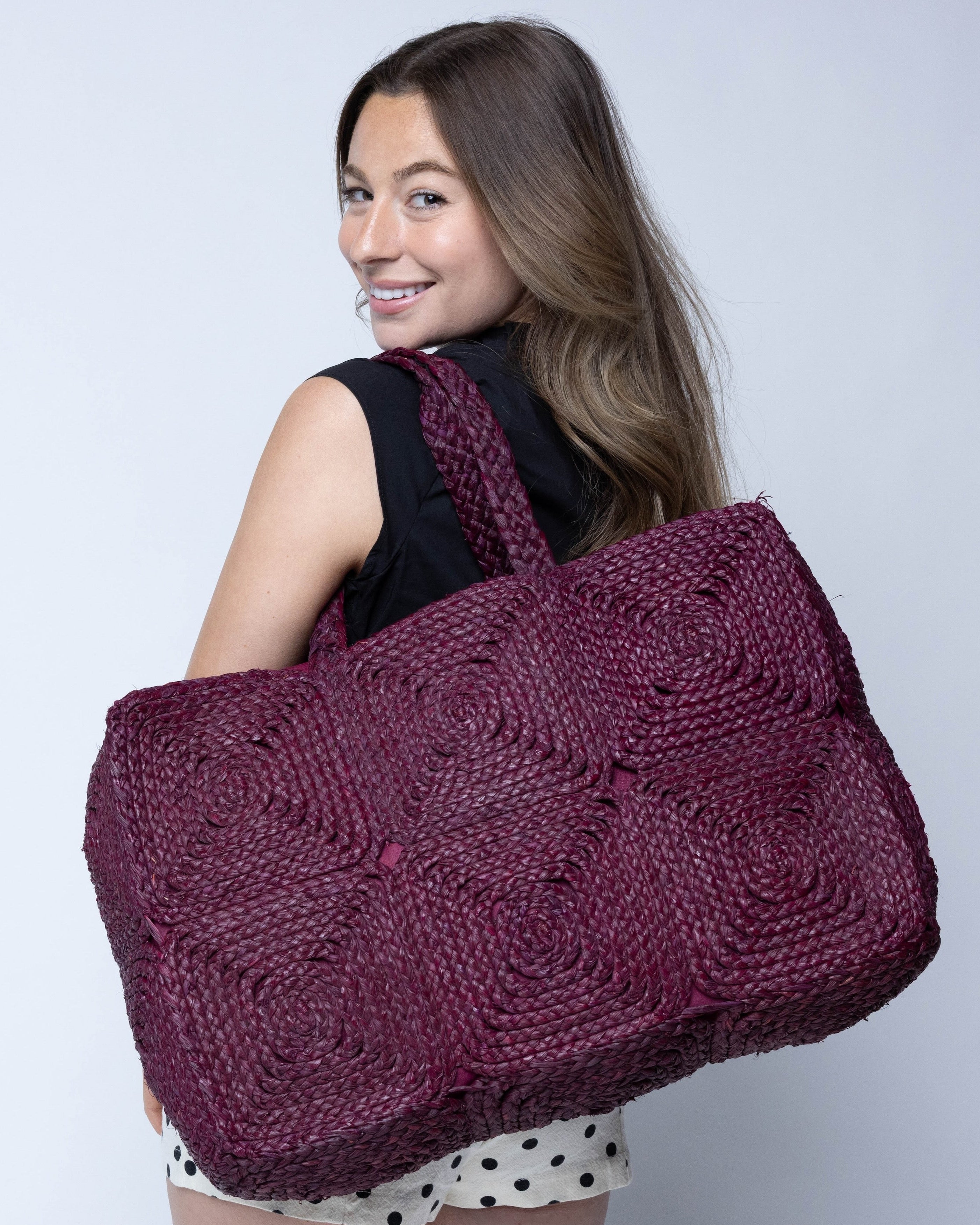Model wearing Gemma Crochet Disc Rectangle Large Straw Basket Handbag handmade braided natural raffia palm fiber in a solid hue of Umberwine burgundy/bordeaux/dark/purple/red with subtle square disc pattern around the outside of the bag in two horizontal bands of squares creating a geometric pattern and matching woven handles beach bag purse - Shebobo