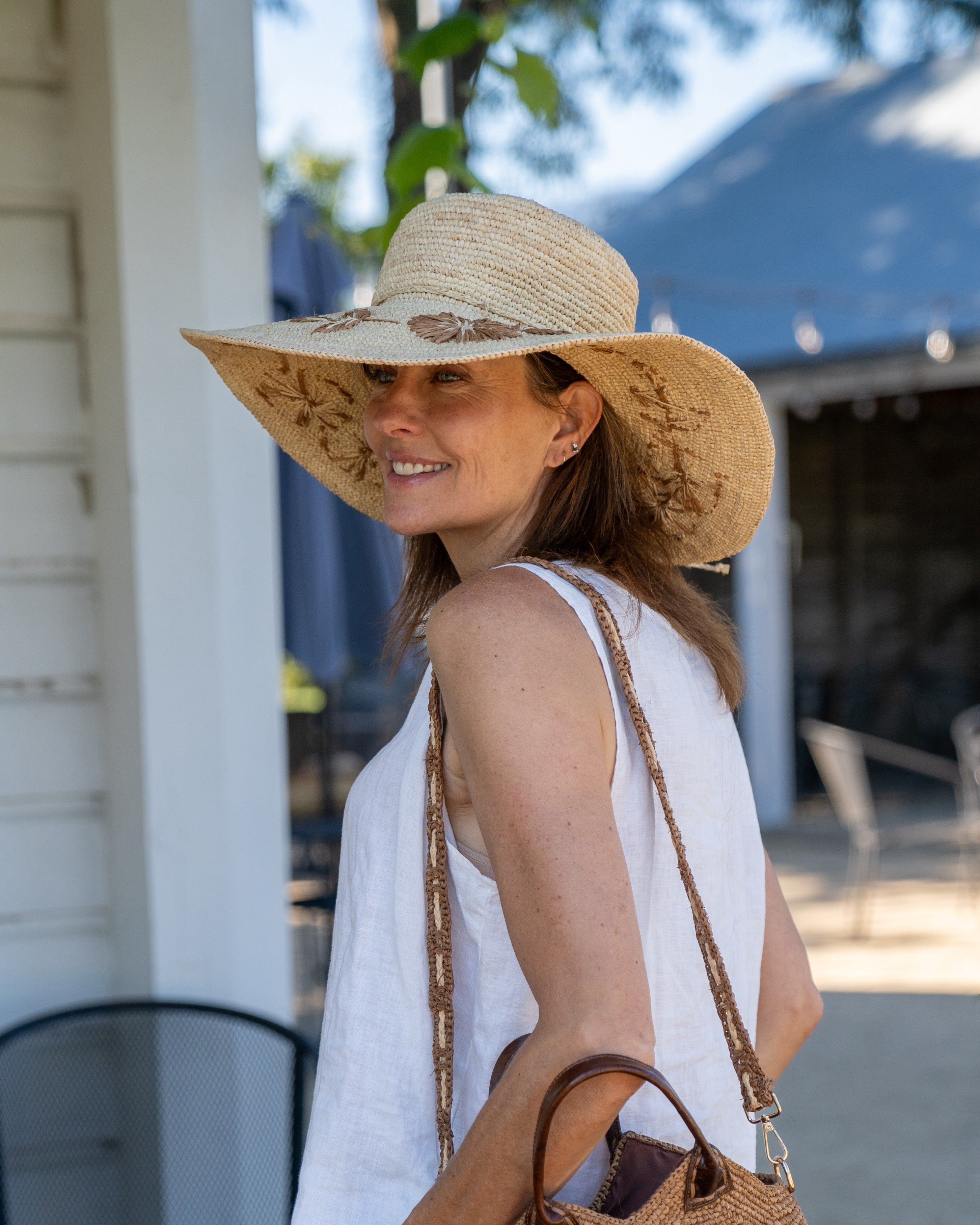Model wearing 5.5" Brim Gigi Natural and Cinnamon Floral Embroidered Crochet Straw Sun Hats handmade crochet natural raffia palm fiber in a solid hue of natural straw color making a concentric pattern from the center top of the flattened crown spiraling out to the edge of the brim with matching adjustable raffia braid hat band and cinnamon dark brown floral pattern with leaves embroidered on the brim - Shebobo