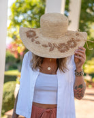 Model wearing 5.5" Brim Gigi Natural and Cinnamon Floral Embroidered Crochet Straw Sun Hats handmade crochet natural raffia palm fiber in a solid hue of natural straw color making a concentric pattern from the center top of the flattened crown spiraling out to the edge of the brim with matching adjustable raffia braid hat band and cinnamon dark brown floral pattern with leaves embroidered on the brim - Shebobo