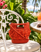 Lifestyle view Hana Mini ConCon Crochet Petite Raffia Straw Basket Handbag Convertible Crossbody Bag with Fringe Purse handmade raffia palm fiber in a solid hue of coral orange/red in a subtle band crochet link texture with horizontal multi tired layers of fringe around the center third of the bag with matching detachable crossbody strap - Shebobo