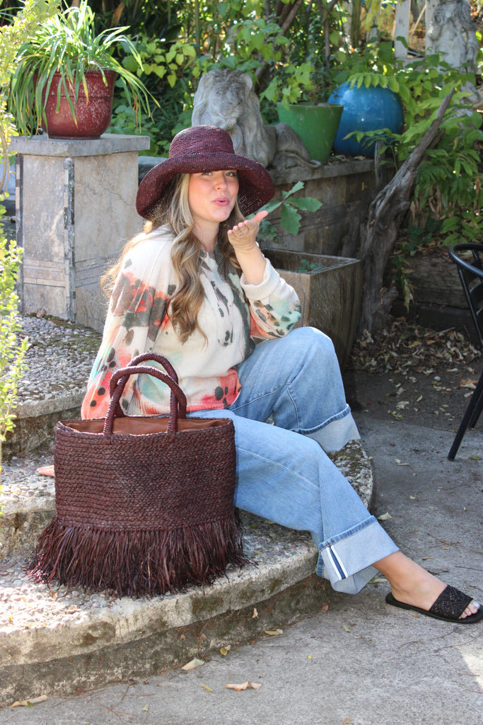 Model wearing Hanalei Braided Straw Basket Shoulder Bag with Fringe handmade braided natural raffia palm fiber in a solid hue of Mocha chocolate/dark/brown with horizontal bands of braids  making a subtle texture and knotted long brushed fringe band around the botom third of the bag and matching looped handles beach bag purse - Shebobo (with Leor 3" Brim Crochet Straw Sun Hats & Lulu Macrame and Crochet Diamond Straw Sandal Shoes)