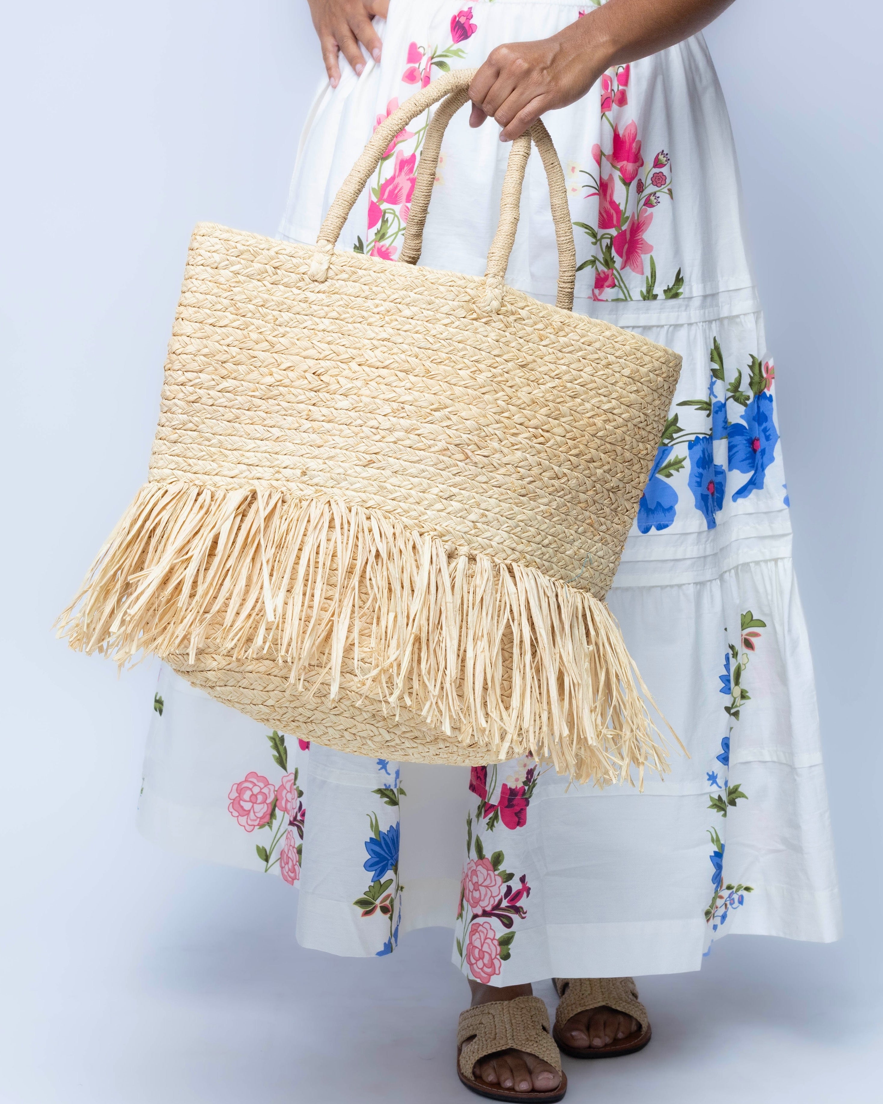 Model wearing Hanalei Braided Straw Basket Shoulder Bag with Fringe handmade braided natural raffia palm fiber in a solid hue of Natural straw color with horizontal bands of braids  making a subtle texture and knotted long brushed fringe band around the botom third of the bag and matching looped handles beach bag purse - Shebobo