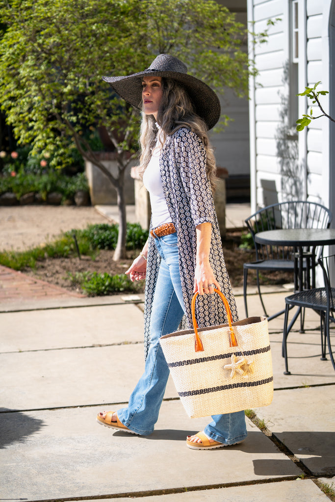 Model wearing Havana Sisal Basket Bag with Tassel Charm Embellishment handmade sisal fiber woven into horizontal wide bands of natural straw color with two evenly spaced narrow bands of black create a stripe pattern with matching crochet starfish charm embellishment and leather handles handbag shoulder bag - Shebobo (with 7" Wide Brim Natasha Woven Straw Sun Hat)