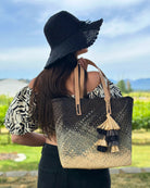 Model wearing Holden Ombre Black Straw Handbag Tassel Charm Embellishment handmade woven raffia dip dyed gradient of black, grey, and natural with matching multicolored, multi-tiered raffia tuft tassel with 4" Brim Genevieve Crochet Straw Sun Hats with Lace Weave Edge Black - Shebobo