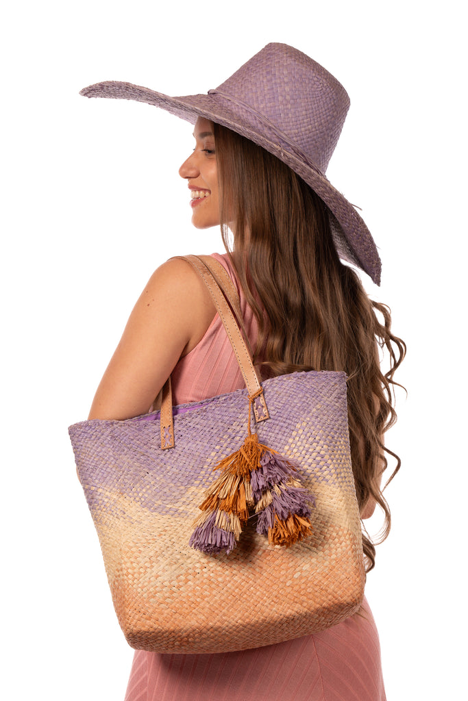 Model wearing Holden Ombre Wisteria Straw Handbag Tassel Charm Embellishment handmade woven raffia dip dyed gradient of wisteria lavender/lilac/light/purple, natural straw color, and blush orange/pink with matching multicolored, multi-tiered raffia tuft tassel - Shebobo (with 7" Wide Brim Natasha Wisteria Woven Straw Sun Hat)