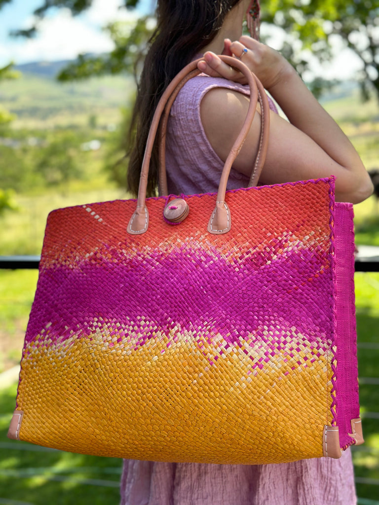 Model wearing Holden Fuchsia Ombre Zafran Large Straw Beach Bags handmade dip dyed raffia woven into a three part horizontal ombre pattern of coral orange/red on the top, fuchsia pink in the middle and both sides, and saffron yellow on the bottom with cross stitch edging, wooden button, and leather handles - Shebobo