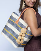 Model wearing Imperial Handbag natural sisal fiber in natural straw color, grey, and saffron yellow horizontal multi-width stripe pattern handmade woven basket with raffia waterfall pompom charm embellishment - Shebobo
