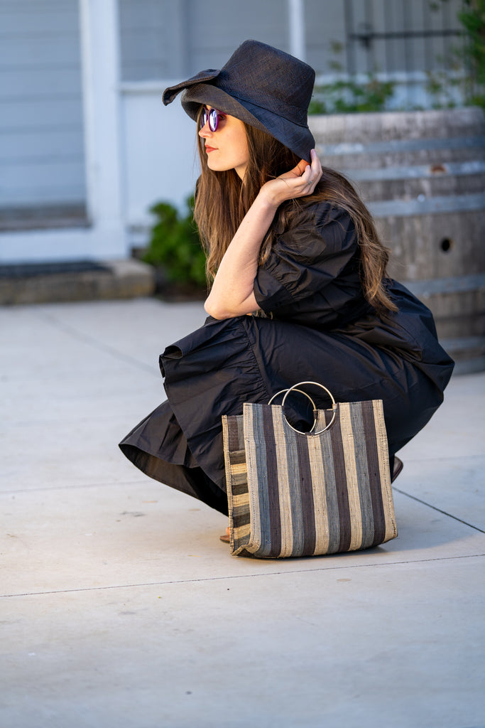 Model wearing Black Swirl JoJo Woven Straw Handbag with Metal Ring Handle handmade natural raffia palm fiber in a stripe pattern of natural straw color, grey, and black with matching woven thread texture and metallic circular metal ring handle - Shebobo (with 3" Brim Cara Bucket Straw Sun Hats with Big Bow)