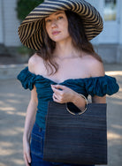 Model wearing JoJo Woven Straw Handbag with Metal Ring Handle handmade natural raffia palm fiber in a solid hue of Black with matching woven thread texture and metallic circular metal ring handle - Shebobo (with Monica 5" & 7" Wide Brim Stripe Packable Straw Sun Hats)