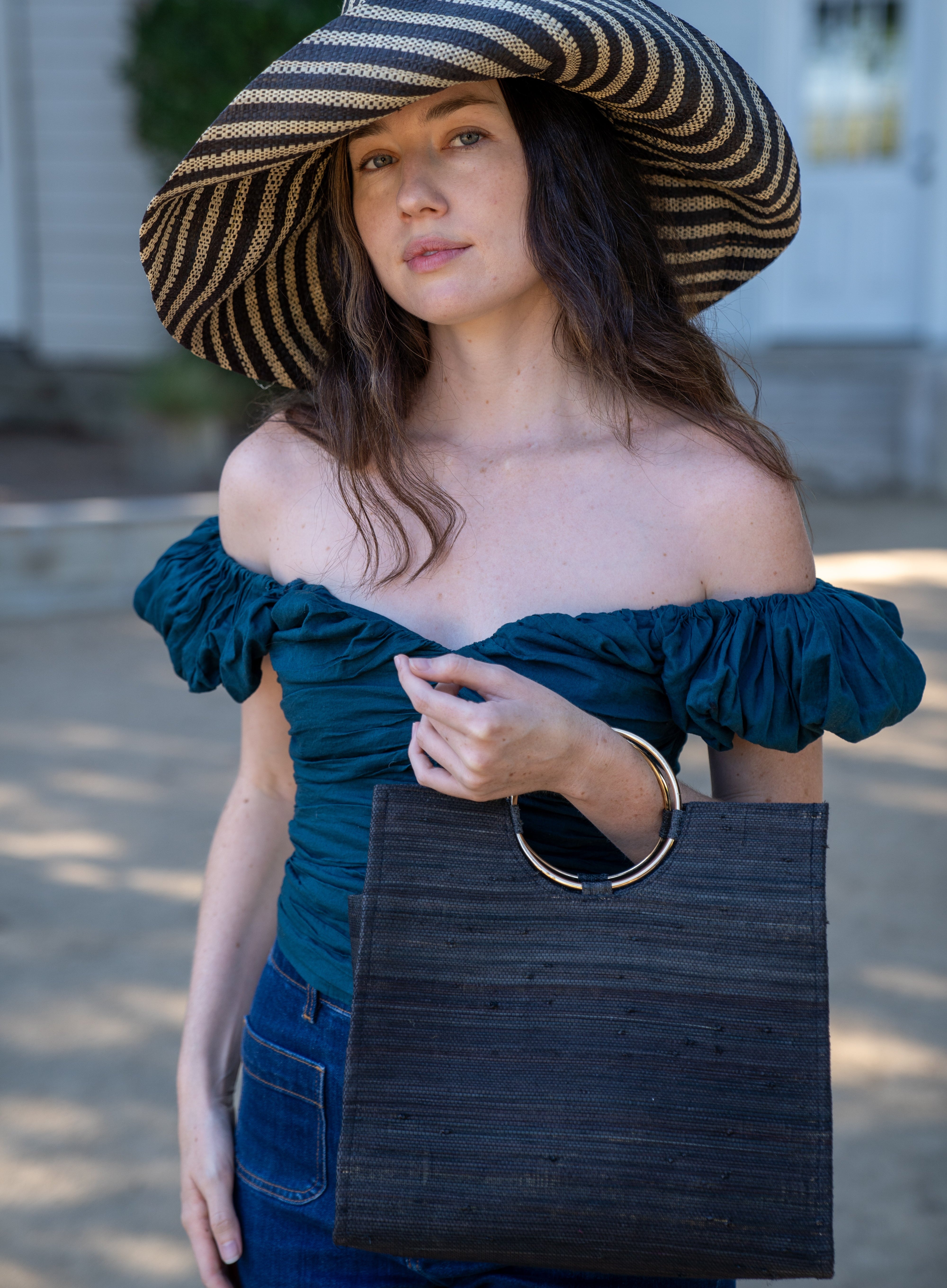 Model wearing JoJo Woven Straw Handbag with Metal Ring Handle handmade natural raffia palm fiber in a solid hue of Black with matching woven thread texture and metallic circular metal ring handle - Shebobo (with Monica 5" & 7" Wide Brim Stripe Packable Straw Sun Hats)
