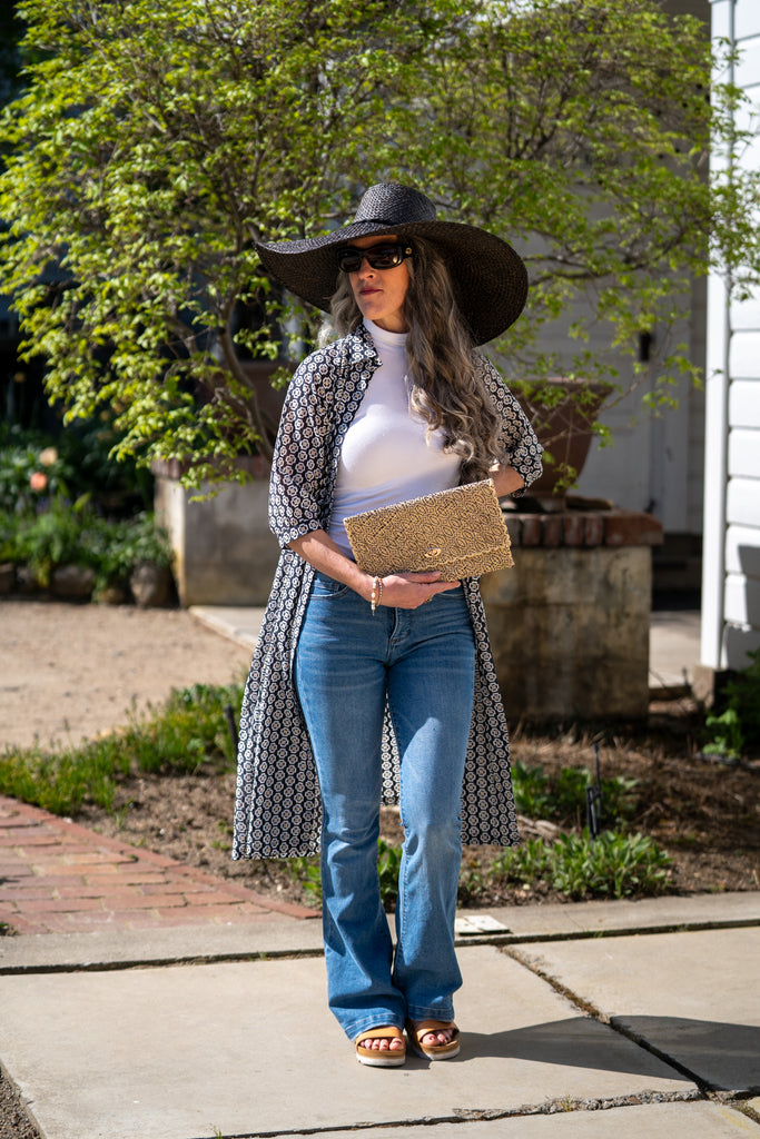 Model wearing Josephine Woven Straw Clutch Purse handmade natural raffia palm fiber in natural straw color woven with contrasting black thread stitching in a subtle diagonal stripe pattern consisting of bands of ovals and circles handbag with crosshatch stitch edging - Shebobo (with 7" Brim Natasha Woven XL Straw Sun Hats)