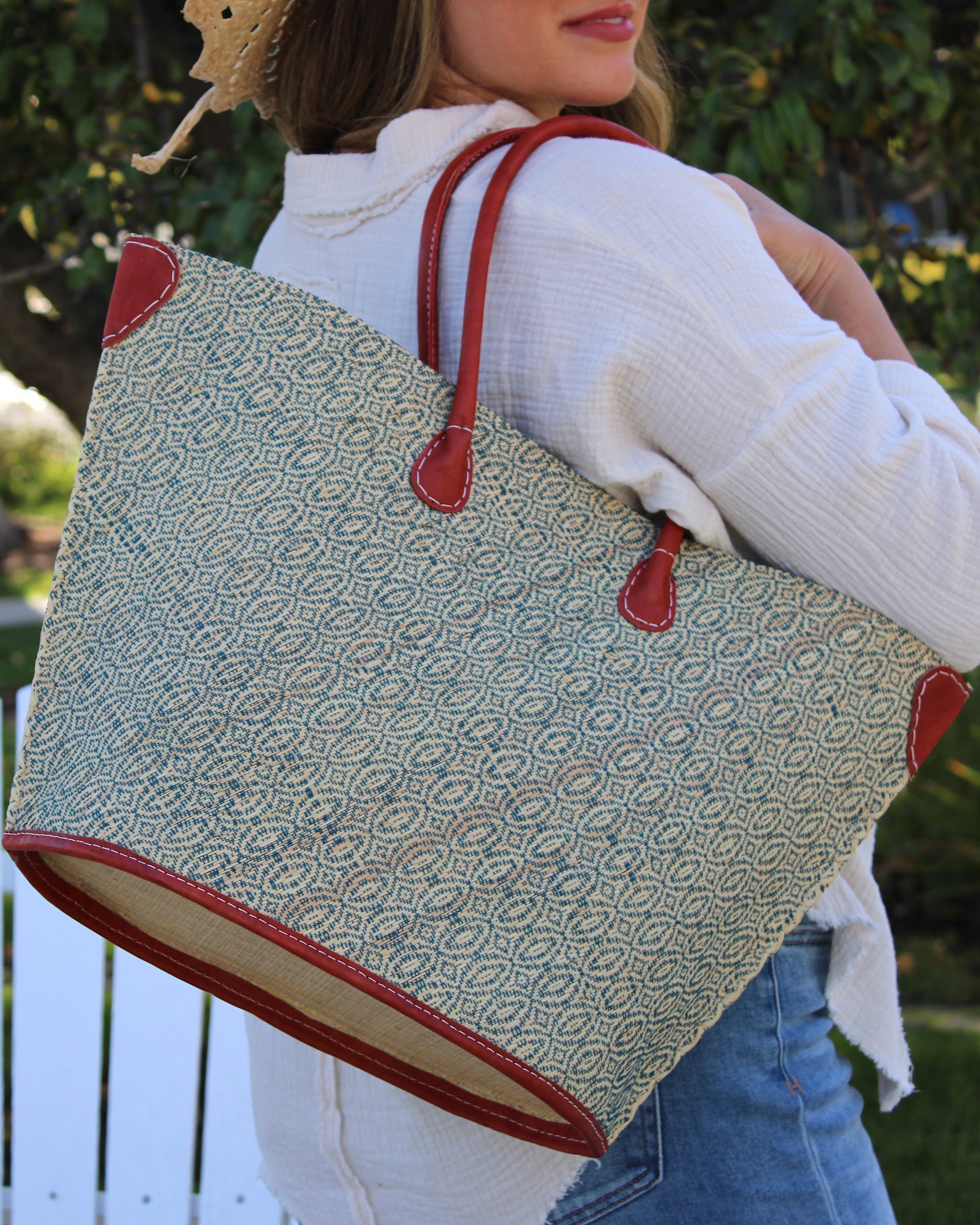 Model wearing Josephine Woven Small Straw Tote Bag handmade natural raffia palm fiber in natural straw color woven with contrasting Turquoise blue/green thread stitching in a subtle diagonal stripe pattern consisting of bands of ovals and circles with natural cross-stitch edge and leather handles beach bag shoulder bag - Shebobo