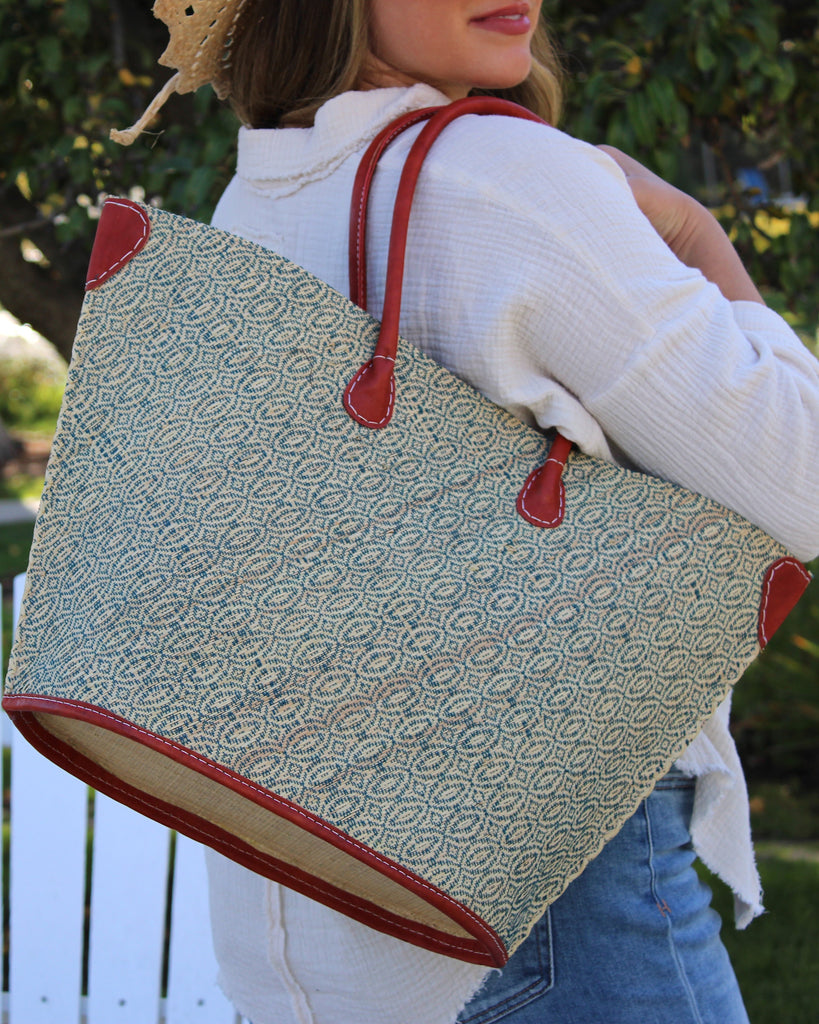 Model wearing Josephine Woven Small Straw Tote Bag handmade natural raffia palm fiber in natural straw color woven with contrasting Turquoise blue/green thread stitching in a subtle diagonal stripe pattern consisting of bands of ovals and circles with natural cross-stitch edge and leather handles beach bag shoulder bag - Shebobo