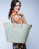 Model wearing Josephine Woven Small Straw Tote Bag handmade natural raffia palm fiber in natural straw color woven with contrasting Turquoise blue/green thread stitching in a subtle diagonal stripe pattern consisting of bands of ovals and circles with natural cross-stitch edge and leather handles beach bag shoulder bag - Shebobo