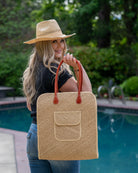 Model wearing Josephine 50's Inspired Woven Straw Handbag handmade natural raffia palm fiber in natural straw color woven with contrasting Cinnamon tobacco/dark/brown thread stitching in a subtle diagonal stripe pattern consisting of bands of ovals and circles with loomed raffia edging around the bag as well as the pocket and flap of the exterior pocket - Shebobo (with Macho Unisex Straw Cowboy Hats with Adjustable Wire Edge)