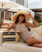 Model wearing xImperial sisal basket natural straw color, black, and grey horizontal multi-width stripe pattern handmade woven natural sisal fiber basket bag with raffia waterfall pompom charm embellishment handbag purse with 5" Wide Brim Natural Woven Kat Straw Sun Hat - Shebobo