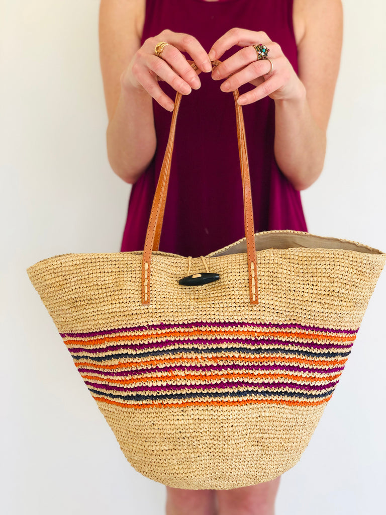 Model wearing Kerry Crochet Bucket Handbag handmade woven raffia multicolor fuchsia pink, coral orange, and navy blue narrow horizontal pinstripe pattern centered within two bands of natural straw color with horn button closure and leather handles purse bag - Shebobo