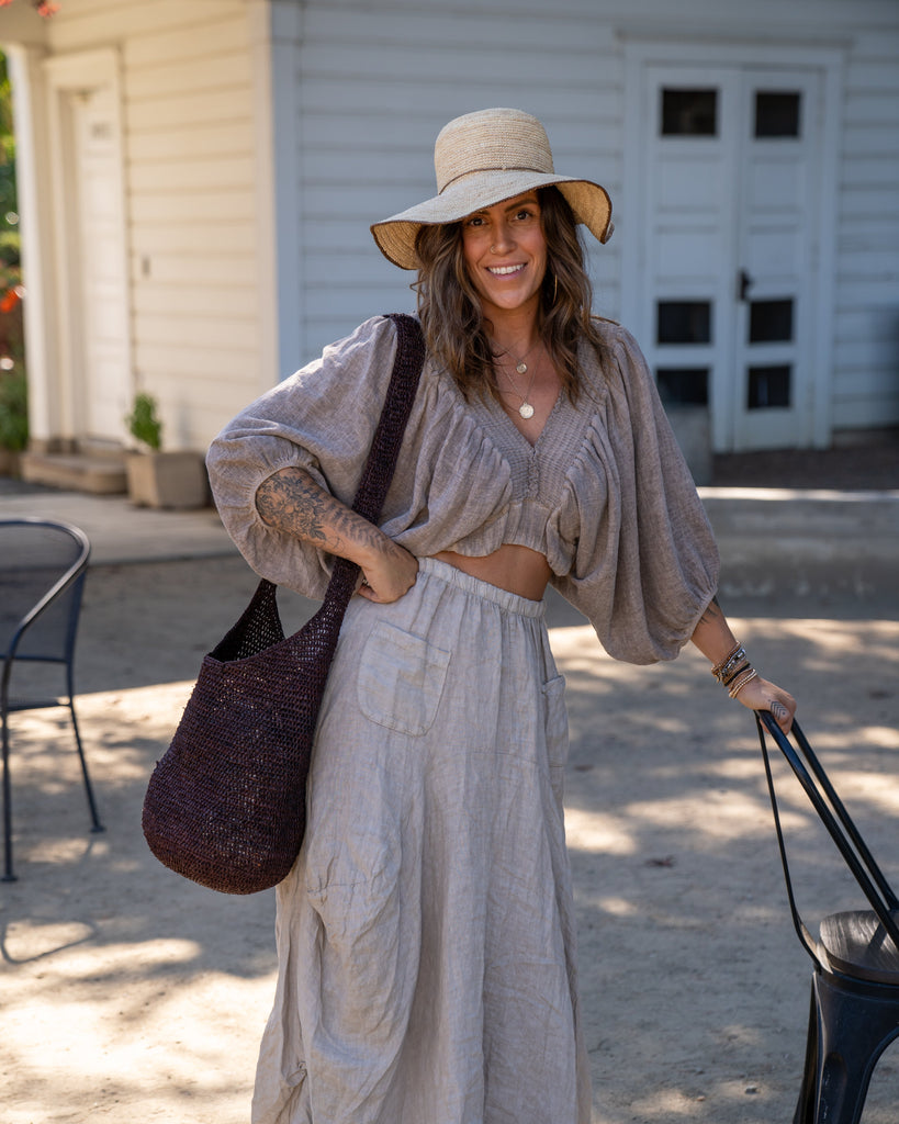Model wearing Model wearing Kiki Crochet Straw Crossbody Bag handmade natural raffia palm fiber in a crochet pattern of subtle linked bands in a solid hue of dark chocolate brown purse - Shebobo (with Rachel 4" Brim Crochet Straw Sun Hats with Colored Edge)