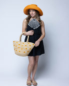 Model wearing Kirra Daisy Flowers Crochet Straw Basket Handbag handmade crochet raffia palm fiber in a solid hue of natural straw color with embroidered round saffron yellow/orange petals and coral orange/red inner dot flower pattern purse with matching crochet handles beach bag purse tote - Shebobo (with Monica Wide Brim Packable Straw Sun Hat)