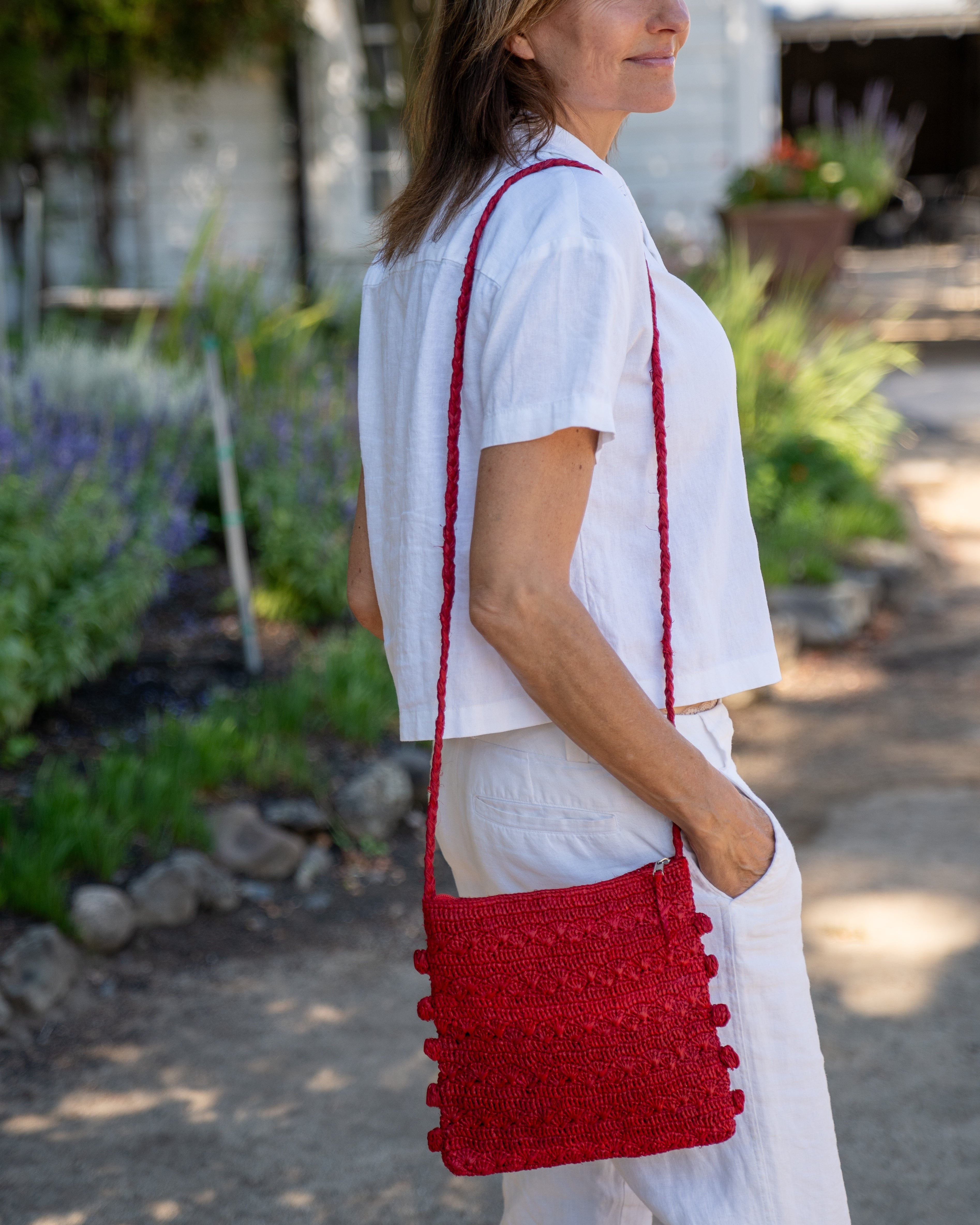 Model wearing Kristen Crochet Straw Crossbody Bag with Nub Edge handmade crochet natural raffia palm fiber in a solid hue of red with two textures of fans and links that create a horizontal stripe pattern across the bag with braided shoulder strap and crochet nub edging along the vertical sides of the purse - Shebobo