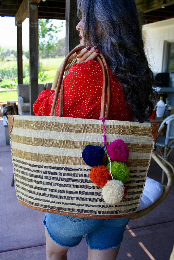Model wearing Laguna handmade loomed raffia handbag straw tote bag cinnamon/tobacco/light brown and natural horizontal stripes of medium width on the top half, and black/chocolate/dark brown and natural narrow width horizontal stripes on the lower half plus leather handles, and a multicolor fuschia pink, navy blue, lime green, orange, natural, and red pompom cluster charm embellishment - Shebobo